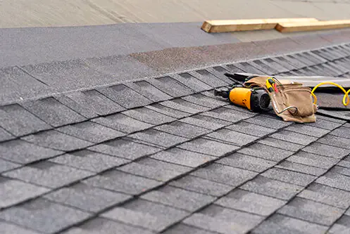 New ridge cap shingles along a roof ridge with a roofing nail gun and tool belt resting on the slope