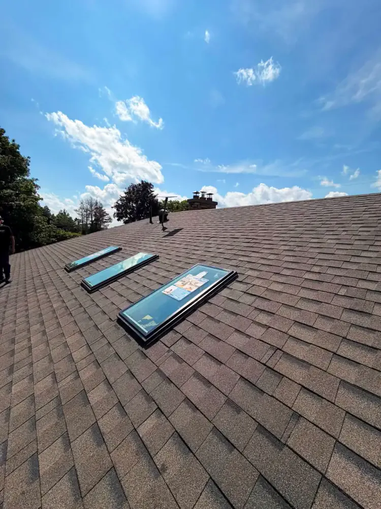Three skylight units laid out on a wide asphalt shingle roof under a blue sky before being fitted